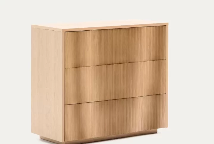 A wooden dresser with a lamp sits beside a neatly made bed in a cozy bedroom setting