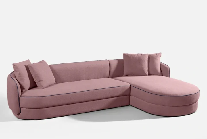 This chic pink sectional sofa features a sleek chaise lounge and a soft rounded design that adds charm to contemporary interiors. Upholstered in plush fabric, it includes three matching cushions and subtle dark piping along the seams for a modern contrast. Ideal for living rooms that embrace playful sophistication, this sofa merges comfort, visual warmth, and minimalism in one standout piece.