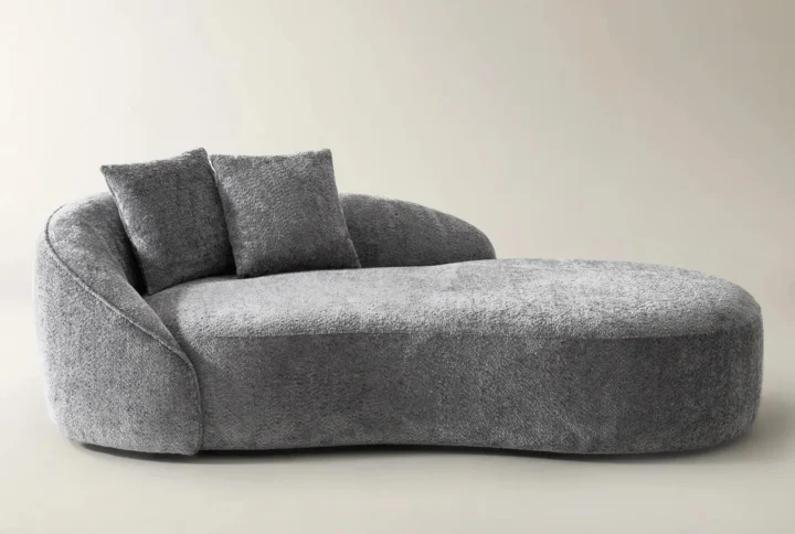 Light gray curved chaise lounge with plush chenille upholstery and matching throw pillows in a modern setting.