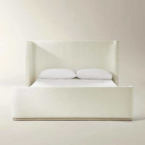 Light beige upholstered bed with curved headboard, white bedding, and wooden base accent in minimalist setting.